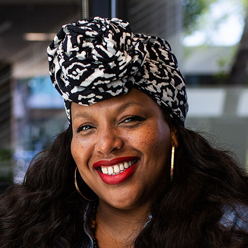 Headshot of Renee Reid. She has a dark skin tone, a black and white knotted headscarf, red lipstick, and gold hoop earrings.